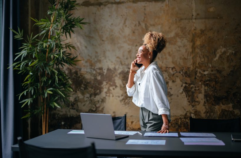 Happy African-American woman speaking on the phone while standing at restaurant desk with a laptop computer and documents.