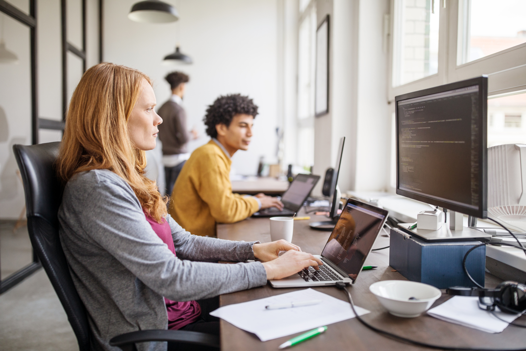 Female professional working on new software program. Businesswoman sitting at her office desk, working on laptop and looking at computer monitor, with colleagues working in background.